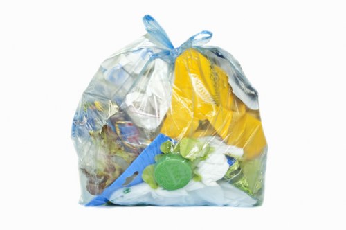 A clear, transparent plastic rubbish bag filled with mixed waste, including yellow and white plastic packaging, a green plastic bottle, and other assorted domestic waste materials. The bag is situated outdoors on a paved surface, with the top gathered and tied into a knot, showing slight bulging indicating it is full. The background is plain and minimal, focusing on the bag as the main object, which is associated with waste disposal and rubbish collection services commonly offered by businesses such as Business Waste Removal Stratford. The textures of the plastic bag appear crinkled and reflective under natural light, with the waste items inside visibly pressed against the sides, emphasizing the contents. The overall scene reflects standard rubbish collection practices in a typical urban or suburban environment, suitable for a service page targeted at local waste removal in Stratford or neighbouring areas.