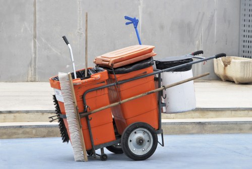 A wheeled orange refuse collection cart filled with cleaning tools and supplies, positioned on a paved surface near a concrete wall. The cart contains a large folded pink plastic sheet, a black item, and a white plastic container, with metal rods and small handles attached. Leaning against the cart are a long-handled broom with black bristles, a push broom with white bristles, and a dustpan with a handle. On the right side, a small beige plastic bin is visible. The background features a grey concrete wall and a section of metallic fencing. The scene suggests a professional rubbish removal or cleaning activity possibly taking place in an outdoor urban or industrial setting, consistent with services provided by Business Waste Removal Stratford in the local area.