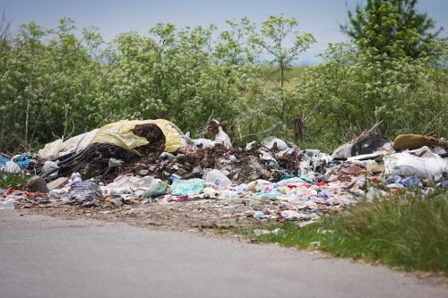 A large accumulation of mixed household and household waste lies spilling onto a gravel roadside area, with various discarded items including plastic bags, cardboard boxes, and fabrics, some partially torn or dirty. The waste heap extends across the foreground, with the debris unevenly piled and extending from the unpaved verge onto the dirt shoulder. Behind the pile, green foliage of bushes and young trees lines the edge of the area, with fresh green leaves indicating a rural or suburban setting, likely near Stratford. The sky above is partly cloudy, illuminating the scene with natural daylight, emphasizing the contrast between the waste and the surrounding natural environment. The image captures a scene that highlights improper waste disposal, which local rubbish removal services such as Business Waste Removal Stratford aim to address, especially near residential or countryside areas within the postcode associated with Stratford.