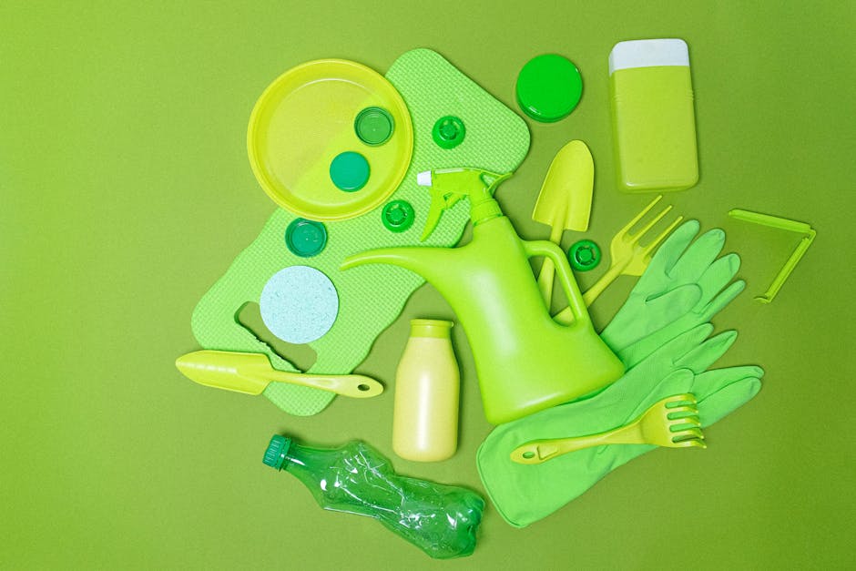 A collection of green household cleaning tools and supplies arranged on a bright green surface. The items include a plastic watering can, a spray bottle, rubber gloves, a small hand rake, a sponge, pl