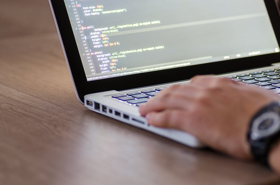 A person with a wristwatch typing on a silver laptop keyboard, which is open on a wooden desk. The laptop screen displays lines of code with syntax highlighting in various colors such as white, yellow